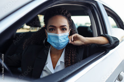 Young business woman fixing and adjusting her medical mask while sitiing in the car behind the steering wheel. Business trips during pandemic, new normal and coronavirus travel safety concept. © Konstantin Zibert