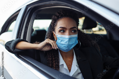 Young business woman fixing and adjusting her medical mask while sitiing in the car behind the steering wheel. Business trips during pandemic, new normal and coronavirus travel safety concept.