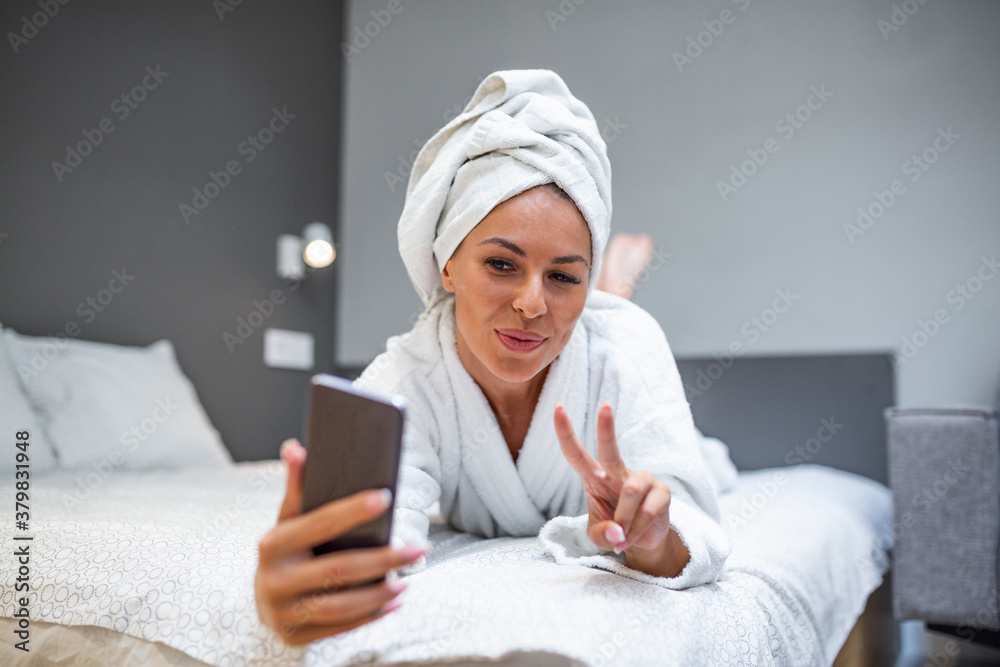Smiling woman lying on bed in bathrobe with mobile phone taking a selfie. Beautiful woman relaxing on the bed after bath and looking at the phone camera taking a selfie showing peace sign Smiling woman lying on bed in bathrobe with mobile phone taking a selfie. Beautiful woman relaxing on the bed after bath and looking at the phone camera taking a selfie showing peace sign