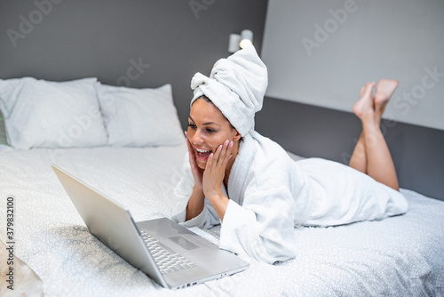 Beautiful young woman in white bathrobe in bedroom at home taking to her friends via video call . Smiling young woman lying on bed in bathrobe relaxing on the bed after bath and looking at her laptop