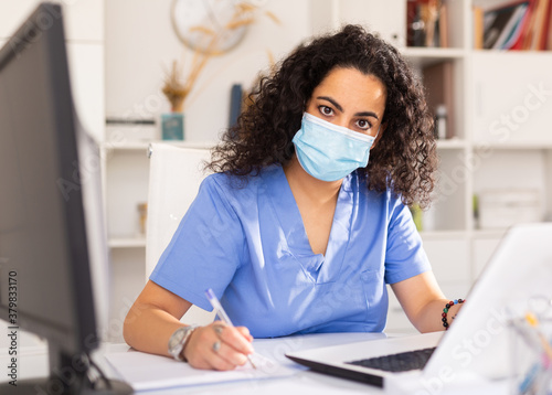 Attentively young female doctor in protective mask working at laptop in her office
