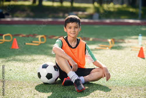 Happy little boy in uniform sitting on soccer field with a ball in front of him after training.