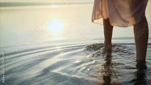 She is wearing a dress. Women's legs in a dress in the water of the lake at sunset. Details of woman's legs. © Ruslan