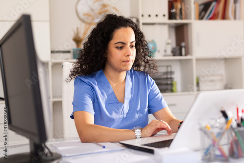 Young woman doctor assistant working in medical office using laptop computer and writing prescription