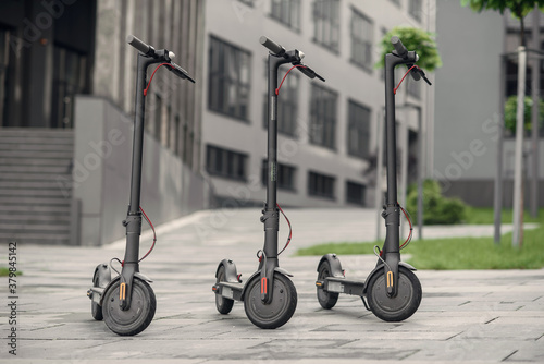 Three modern stylish black electric scooter are parked near big office centre. Electricity and eco friendly concept.
