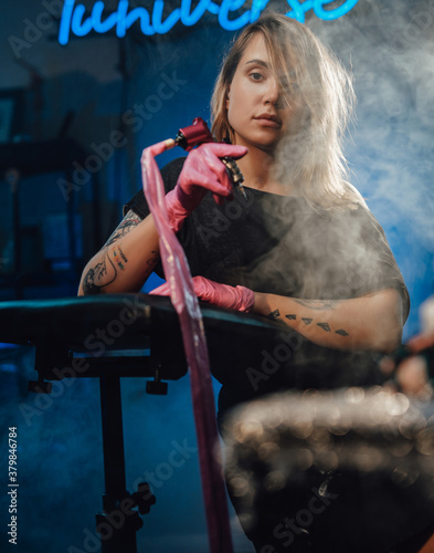 Beautiful female tattoo master with tattoo device on hand posing in her modern and cool workplace in studio with neon title.