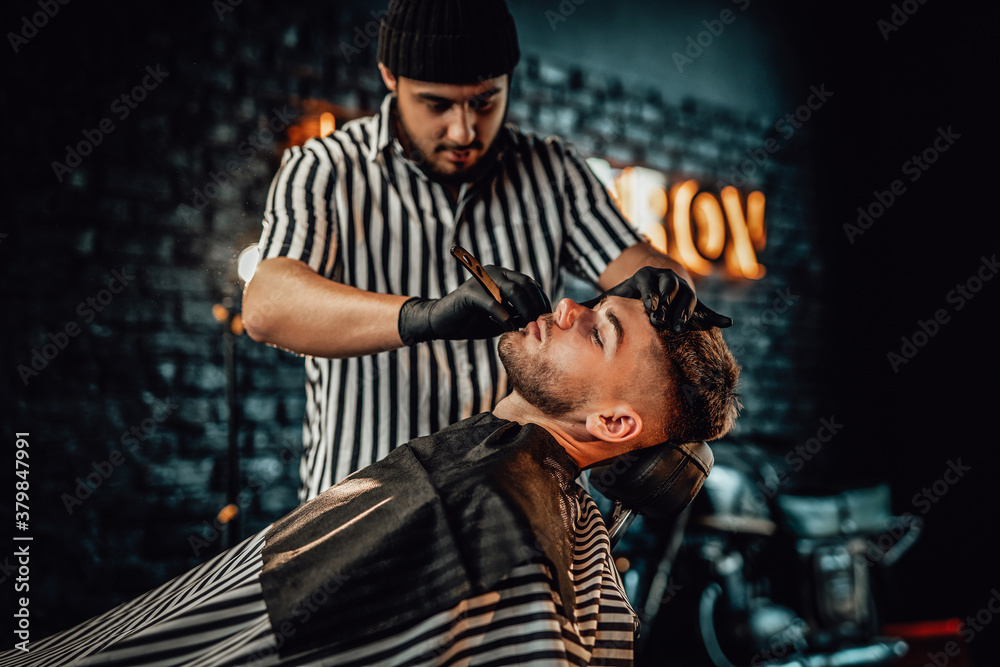 Retro styled barbershop and man is shaving by caucasian barber with black gloves in blurred brickwall background.