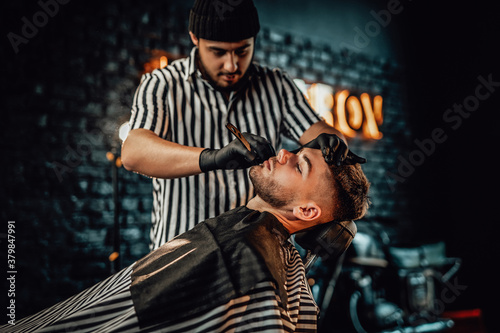 Retro styled barbershop and man is shaving by caucasian barber with black gloves in blurred brickwall background.