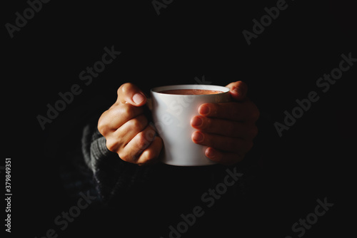 Cup of coffee in women's hands on black background. Woman warming her hands on mug of hot beverage in the dark. Copy space. Minimal style. Low key.