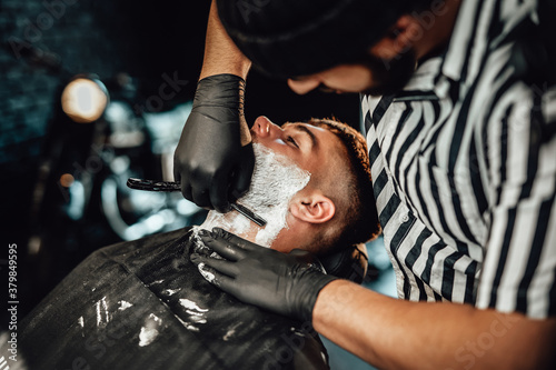 Retro styled barbershop and its worker which carefully cutting his client's beard with modern dark blade.