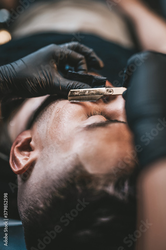 Professional stylist and hairdresser carefully shaving a beard of his client with sharp blade and black gloves.