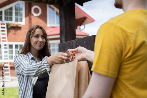 Attractive smiling brunette girl receiving order. uniformed delivery man courier handing paper bag of food to customer by gate of country house. disposable biodegradable bags, recycling concept