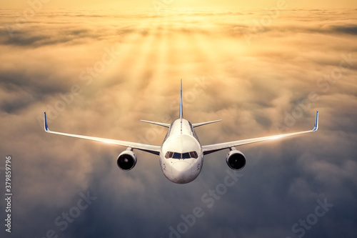 Airplane is flying above the clouds at sunset in summer. Landscape with passenger airplane, low clouds, orange sky. Front view of aircraft. Business travel. Commercial plane. Aerial view. Take off