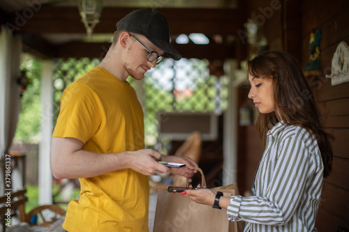 uniformed delivery man handing paper bags of food to Attractive smiling girl. young woman using smartphone to pay for her order. new online payment technologies concept