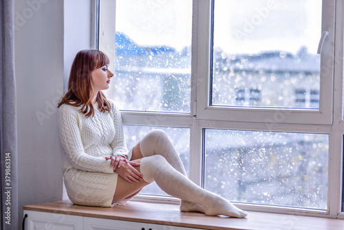 A beautiful young woman in a white knitted sweater dress and long leggings, sits on a wide windowsill at the window. Looking into the distance, sad, loneliness concept. Copy space. White interiors Free Download A beautiful young woman in a white knitted sweater dress and long leggings, sits on a wide windowsill at the window. Looking into the distance, sad, loneliness concept. Copy space. White interiors - GFXLoad