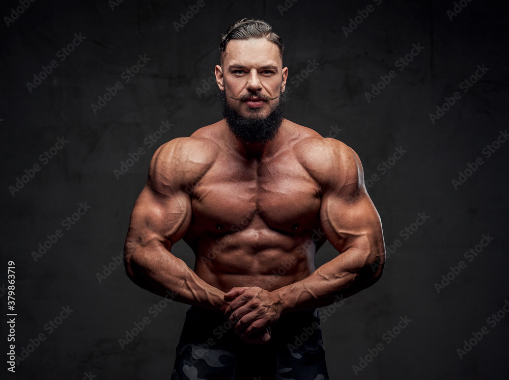 Self confident and aggressive military bodybuilder with naked torso and with black beard posing in gray background.
