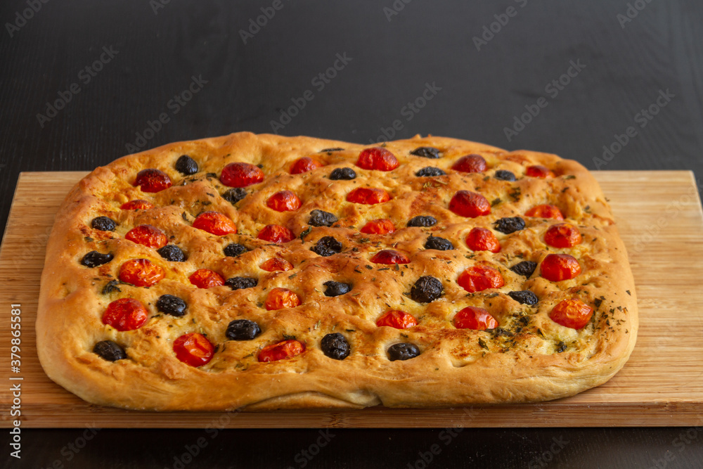 Freshly prepared focaccio with tomatoes and olives lies on a cutting board and stands on a black table.