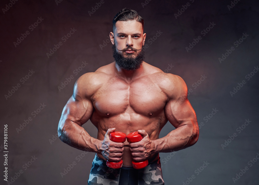 Military bodybuilder with beard holding dumbells and posing in dark gradient background.