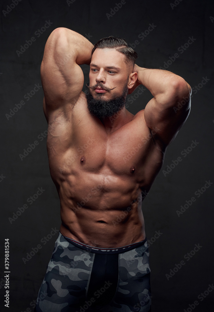 Brutal military athlete with naked torso and with beard and mustache posing in dark gray background.