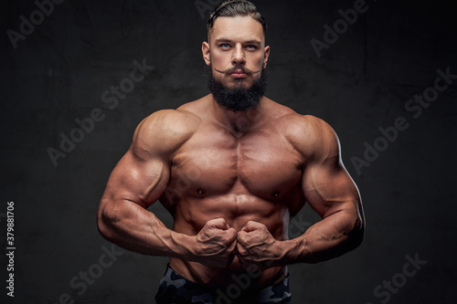 Brutal military athlete with naked torso and with beard and mustache posing in dark gray background. © Fxquadro