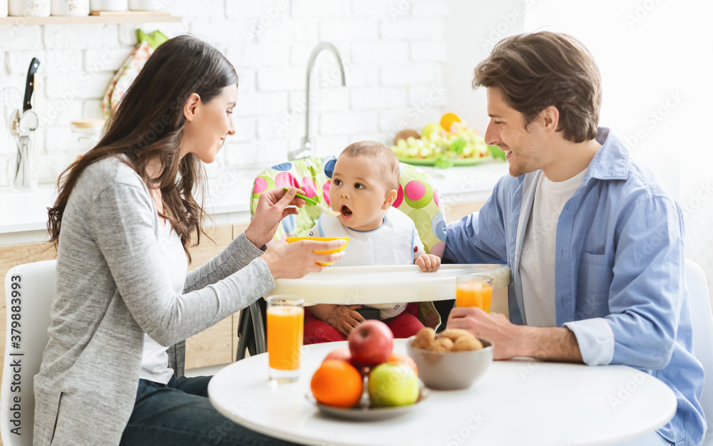 Happy parents feeding baby son at home kitchen