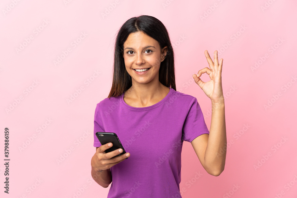 Caucasian girl isolated on pink background using mobile phone and doing OK sign Caucasian girl isolated on pink background using mobile phone and doing OK sign