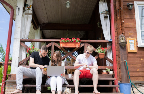 Group Of Cheerful Friends with york dog sit on terrace of wooden house with laptop, smartphone, smart watch communicating in social network. Relax at countryside summer cottage. modern gadgets concept