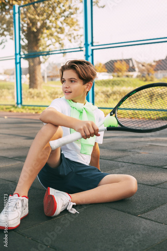 Portrait of a beautiful teenage boy with a tennis racket in his hands. A tennis player sits on the court and rests after training. Sport, sportsman