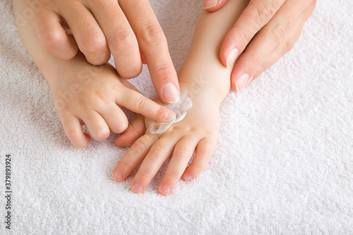 Young mother and baby fingers together applying moisturizing cream on baby hand on white towel. Care about children clean and soft body skin. Front view. Closeup.