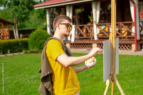 Young attractive man in trendy sunglasses artist painting picture on easel outside on loan near country house  outdoors relaxing activity  creation  inspiration  summer weekend recreation concept
