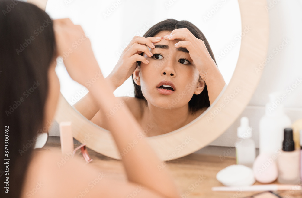 Upset asian young woman checking for pimples, looking at mirror Upset asian young woman checking for pimples, looking at mirror