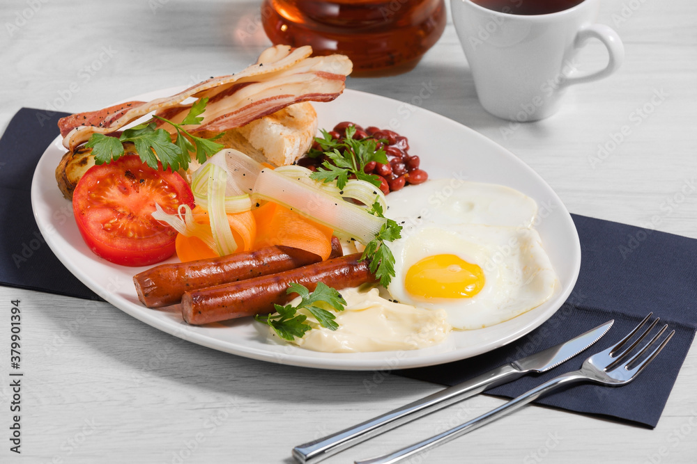Breakfast of grilled sausages, crispy bacon with ciabatta, barbecue beans, fried eggs, grilled and fresh vegetables and cream cheese are on a white plate and a light background. Concept healthy breakf Breakfast of grilled sausages, crispy bacon with ciabatta, barbecue beans, fried eggs, grilled and fresh vegetables and cream cheese are on a white plate and a light background. Concept healthy breakf
