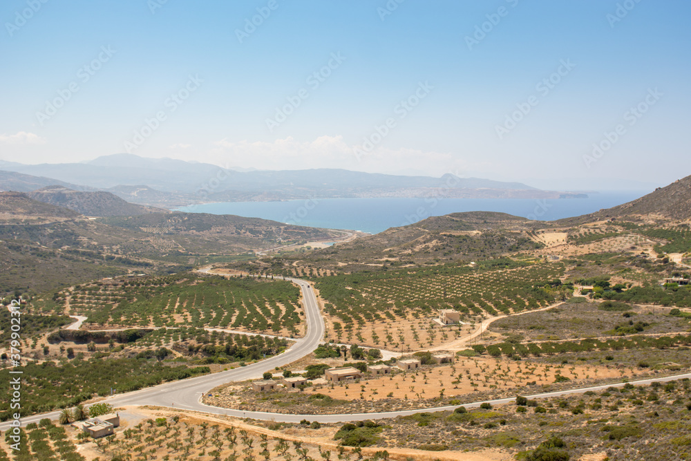 Aerial shot of a road going through fields. Sea and hills in the background. 