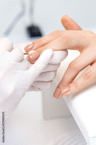 Manicure master in protective gloves applying beige nail polish on female nails in beauty salon