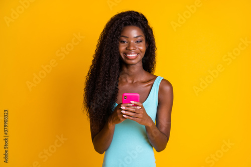 Photo portrait of happy african american woman holding pink phone in two hands smiling wearing blue singlet isolated on vivid yellow colored background