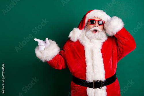 Photo of retired grandpa grey beard open mouth hands head direct finger empty space sale wear red santa x-mas costume coat gloves sunglass belt headwear isolated green color background photo