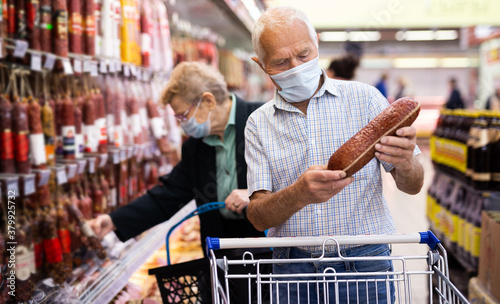 mature european man in mask and gloves with covid protection picks up salami in meat section of supermarket