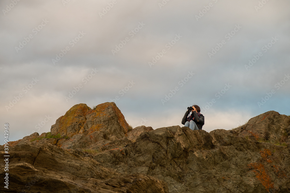 Young woman sitting on a cliff and looking through binoculars in the Bic national park, Canada