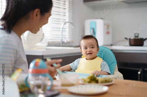 Young mother provide food and fruit puree for lunch to her baby son seat on high chair in kitchen.