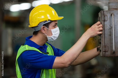 Asian male worker working with machine in factory industry while wearing safety uniform, hardhat and protective face mask