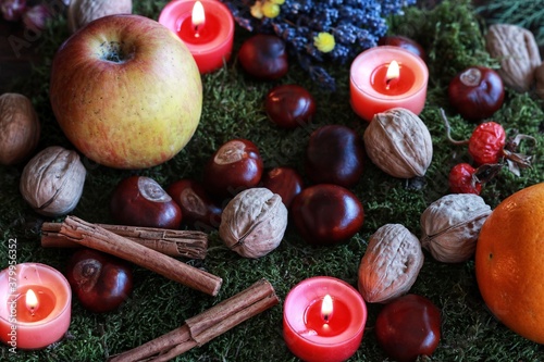 Colorful esoteric autumn fall background filled with September harvest and nature finds. Apple, orange, cinnamon sticks, walnuts, chestnuts and orange colored burning candles on green moss background photo