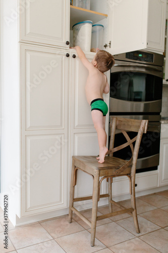 Cute young boy standing on a chair in kitchen looking for treats in cubboard photo