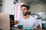 Serious young bearded man browsing smartphone while drinking coffee at table near laptop in cafe