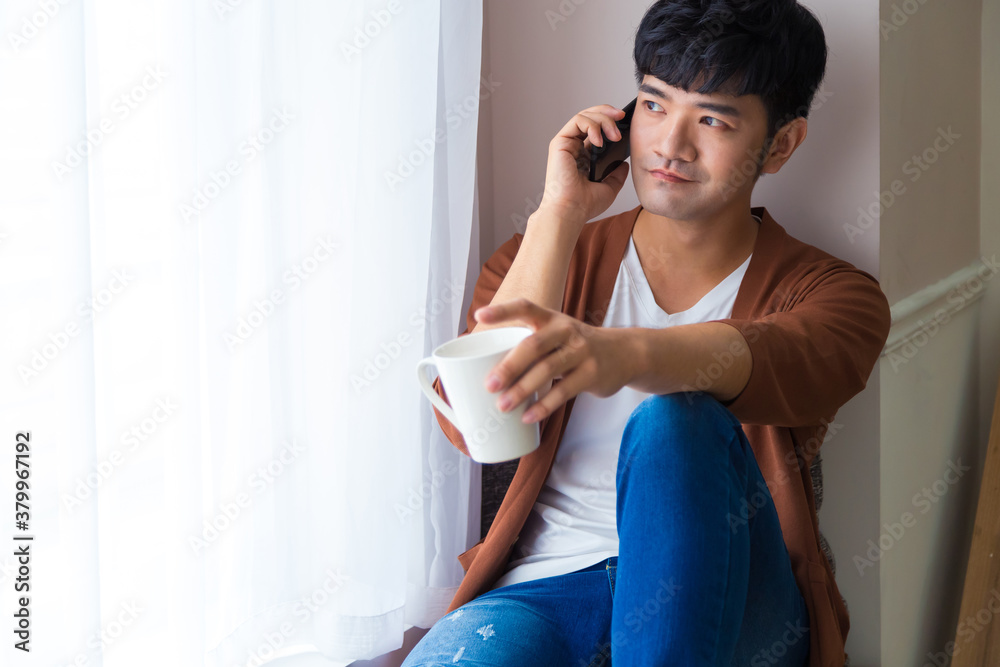 Handsome man is siting near window with cup of coffee and smart phone in hands. Talking on mobile phone. Working at home concept. Handsome man is siting near window with cup of coffee and smart phone in hands. Talking on mobile phone. Working at home concept.