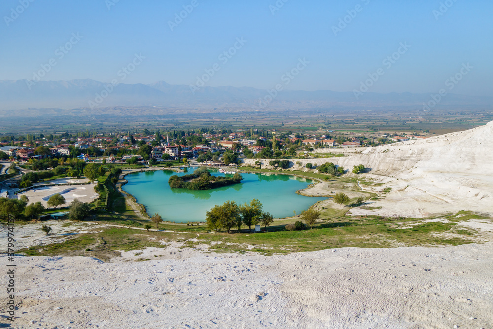 Panoramic view onto lake near foot of famous travertines of Pamukkale (Turkey) & town itself. Bright green & blue color of water formed by mineral water from upper hot spring