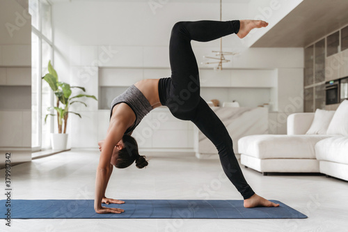 Young attractive fit woman in sportswear practicing yoga, gymnastic, fitness at home, Urdhva Dhanurasana, stretching her back and legs. Indoor full length shot. Wellness and healthy lifestyle concept