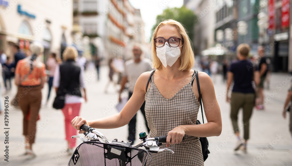 Woman walking by her bicycle on pedestrian city street wearing medical face mask in public to prevent spreading of corona virus. New normal during covid epidemic. Social responsibility. - obrazy, fototapety, plakaty 