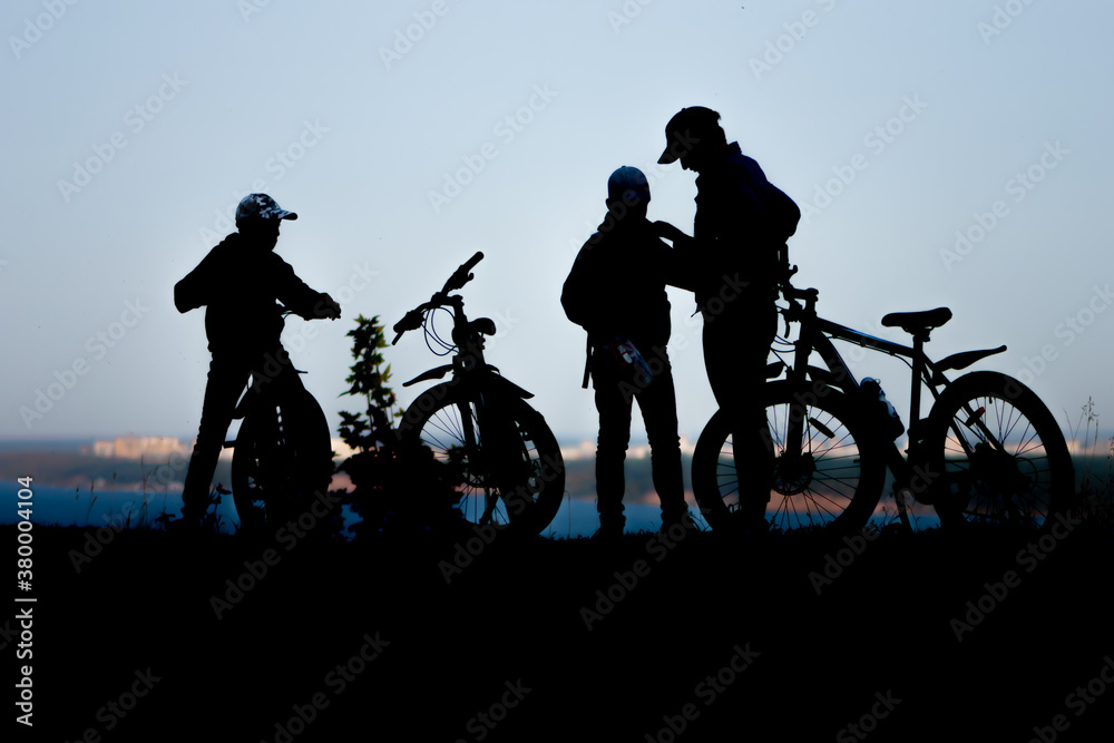 Obraz premium Silhouettes of figures of a father with two sons on a Bicycle ride in the Park by the river. The concept of a healthy lifestyle in the family.
