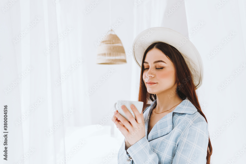 Beautiful young stylish woman in business suit and white hat standing near bed and drink coffee in sunny morning.