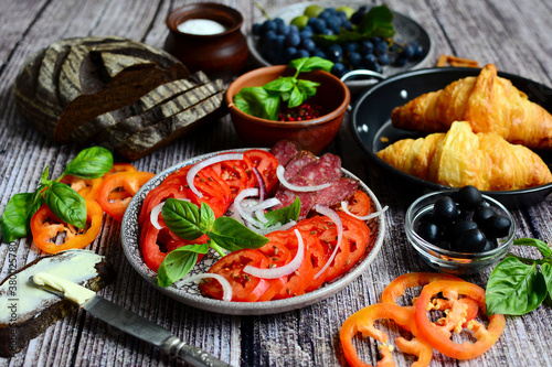 Tomatoes, croissants, fruits, olives and bread on serving plates on a wooden background.